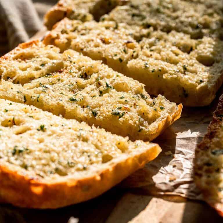 Sliced garlic bread with herbs and melted butter on wood, sunlight emphasizing its golden, crispy texture.