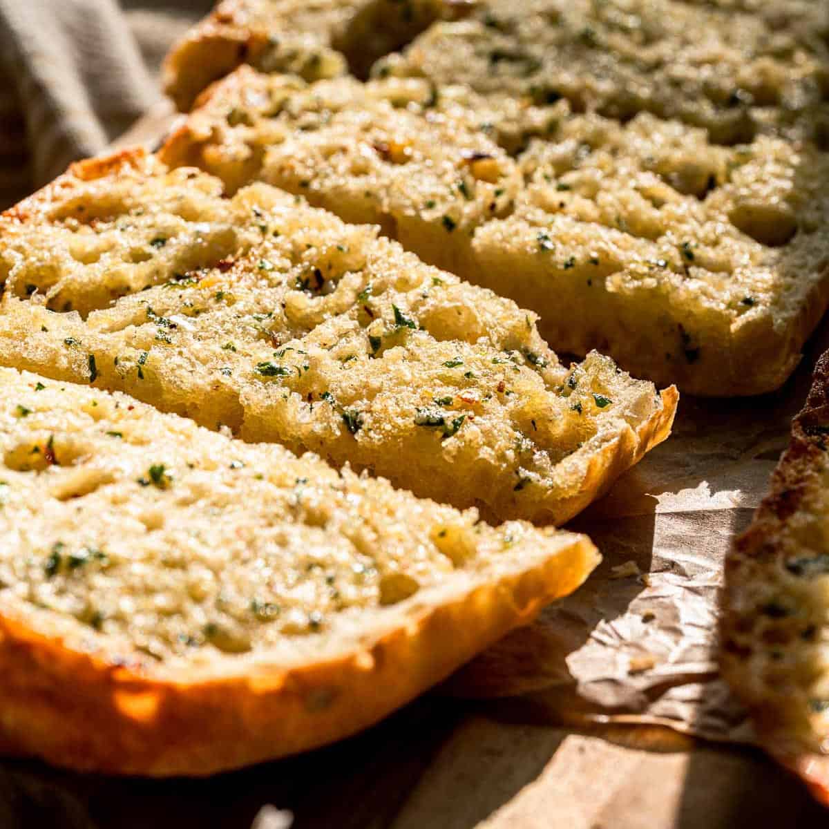 Sliced garlic bread with herbs and melted butter on wood, sunlight emphasizing its golden, crispy texture.