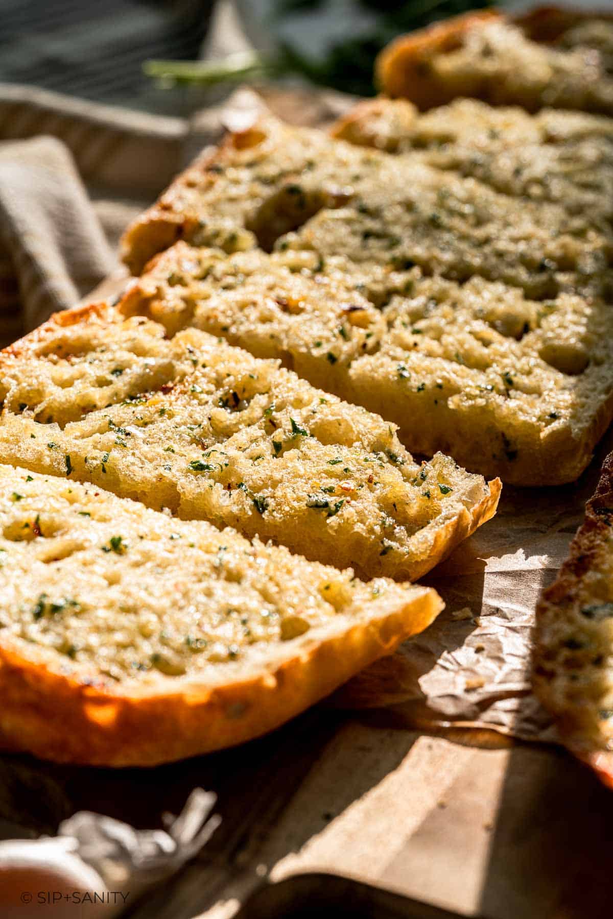 Garlic bread slices with herbs and melted butter on parchment paper in warm natural light.
