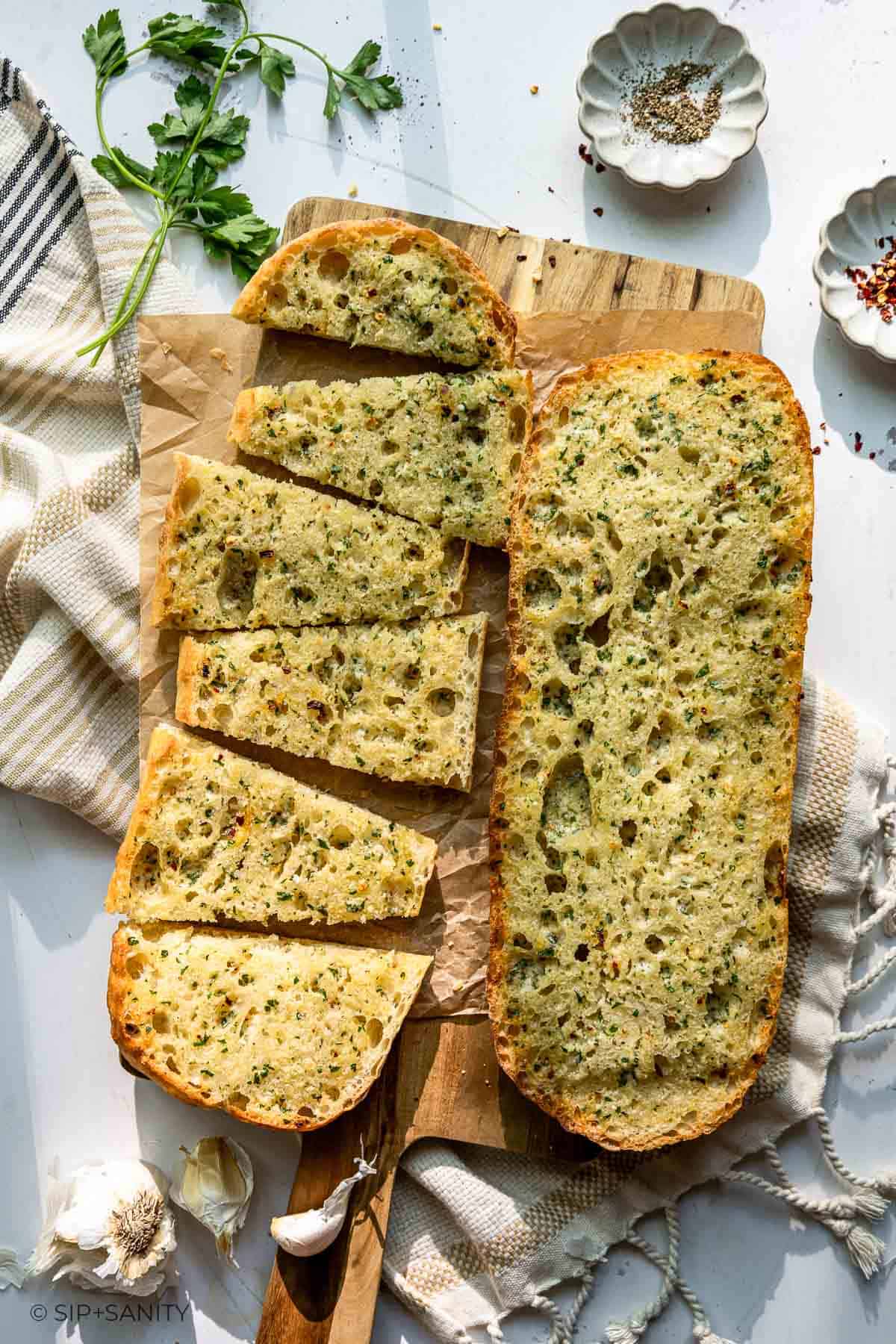 Herb-topped ciabatta garlic bread slices on a wooden board with garlic, parsley, a striped towel, and spice bowls nearby.