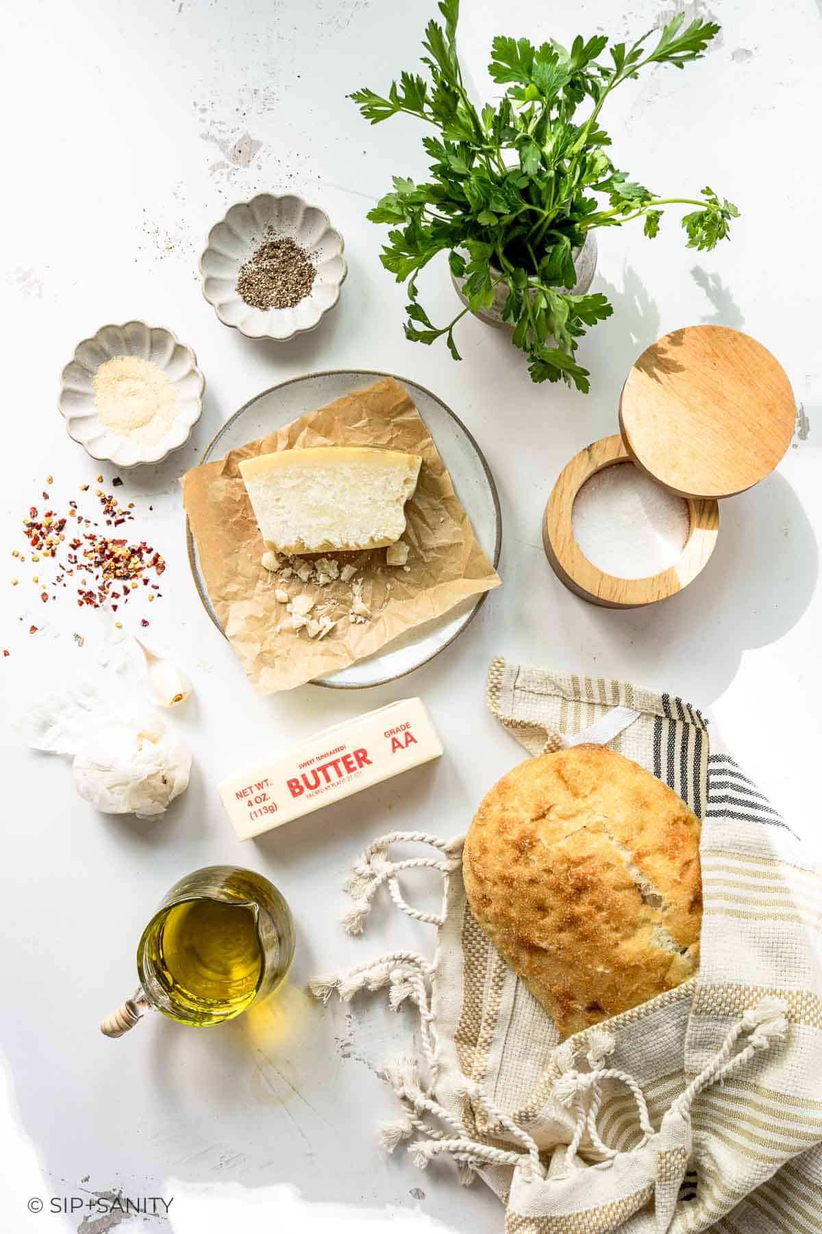 Parmesan, butter, bread, olive oil, garlic, parsley, salt, pepper, red pepper flakes on white with a striped towel.