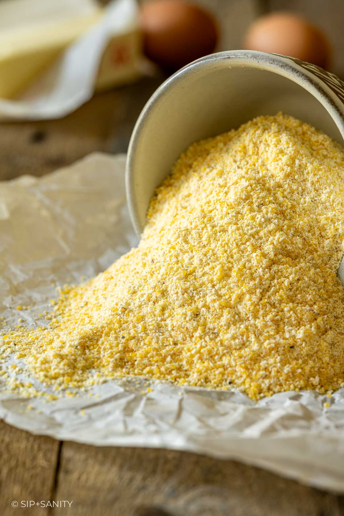Cornmeal spills from a ceramic bowl onto parchment on wood; eggs and a stick of butter are blurred in the background.