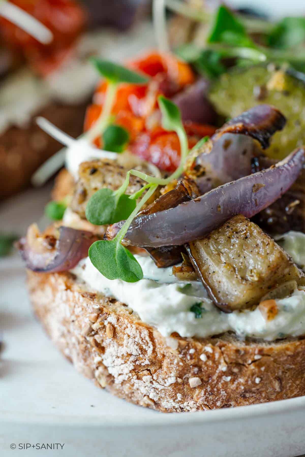 Whole grain bread with cottage cheese, roasted eggplant, red onion, microgreens, and colorful veggies in close-up.