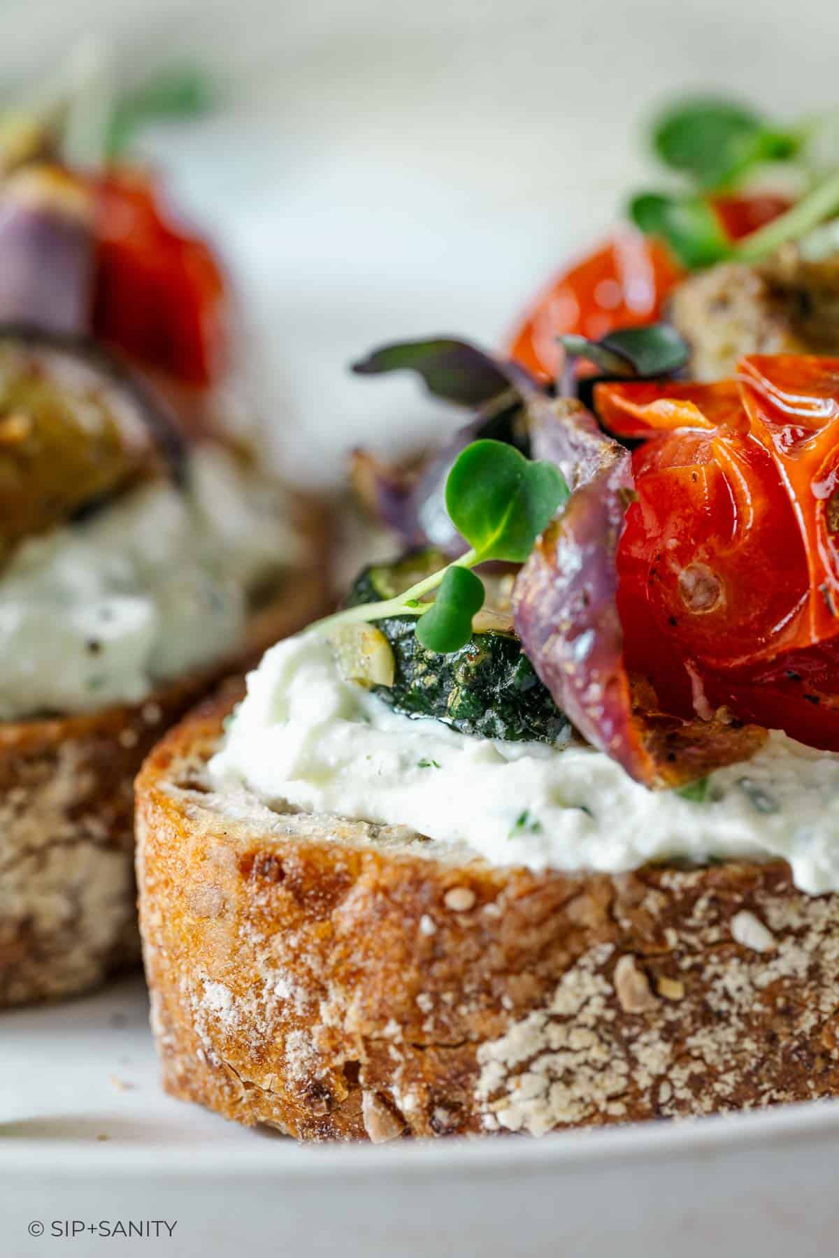 Toasted bread with cottage cheese, roasted cherry tomatoes, red onions, greens, and seasoning on a white surface.