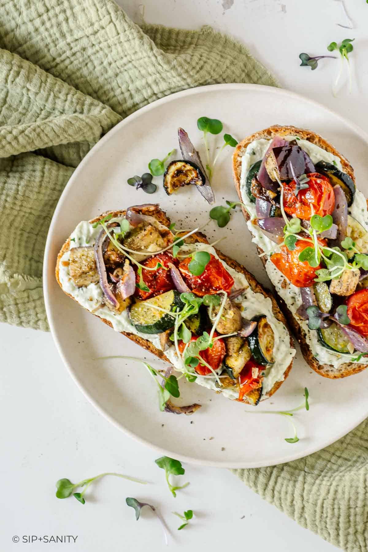 Two toast slices with roasted veggies, microgreens, and creamy cottage cheese on a white plate, green cloth beneath.