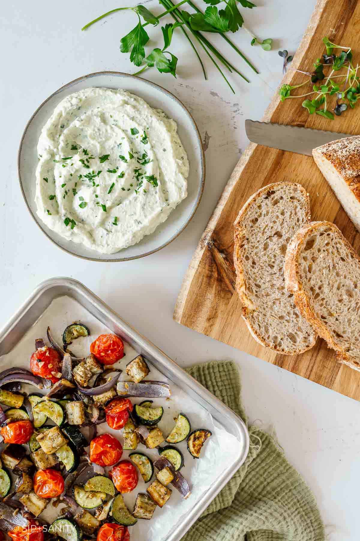 Creamy cottage cheese with herbs, bread slices, roasted zucchini, eggplant, cherry tomatoes, and fresh herbs on white surface.