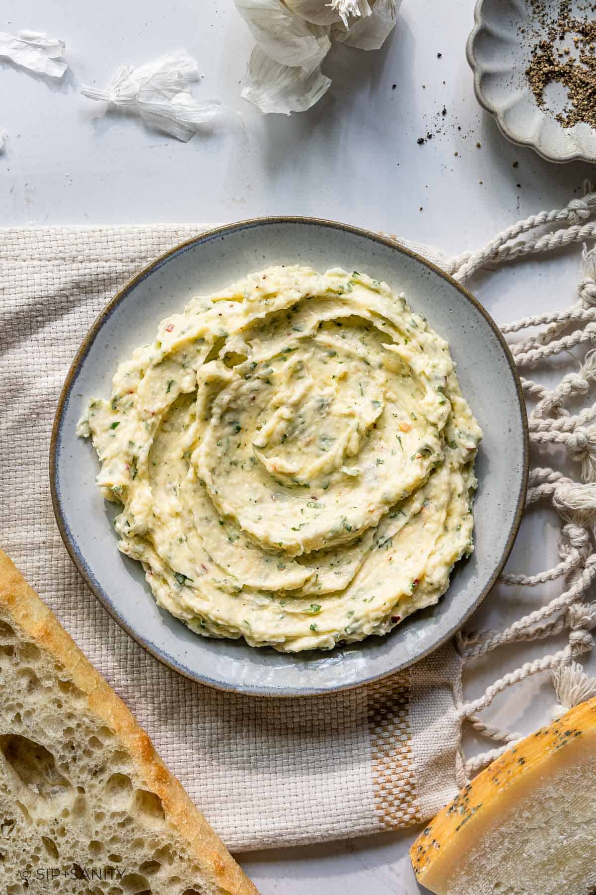 Herb garlic butter with green flecks on a gray plate, beside bread slices, garlic cloves, and black pepper on a towel.