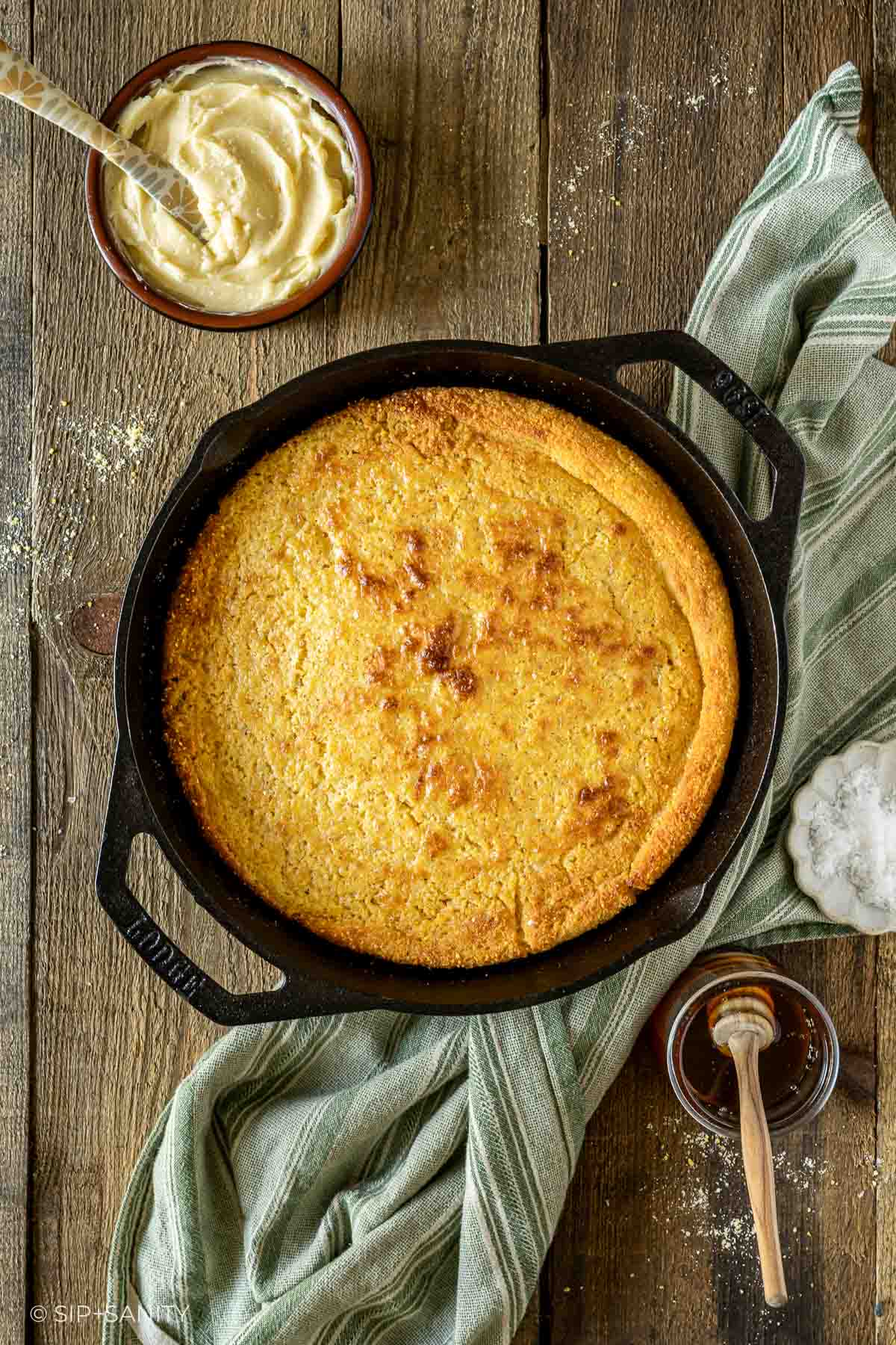 Cornbread in a skillet on a wooden table with whipped butter, striped cloth, and honey jar with dipper.