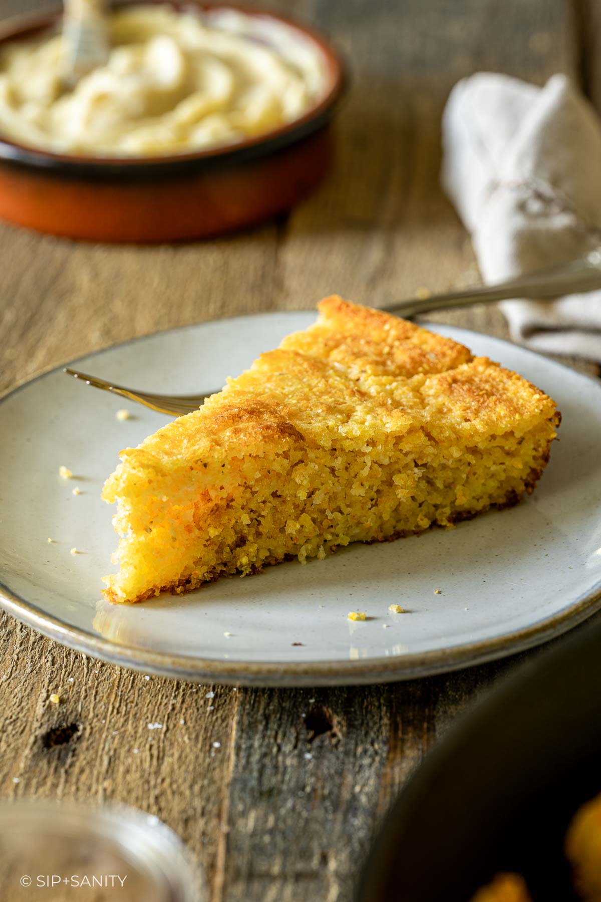 A golden slice of cornbread on a gray plate with a fork on a rustic table; buttery spread blurred in the background.