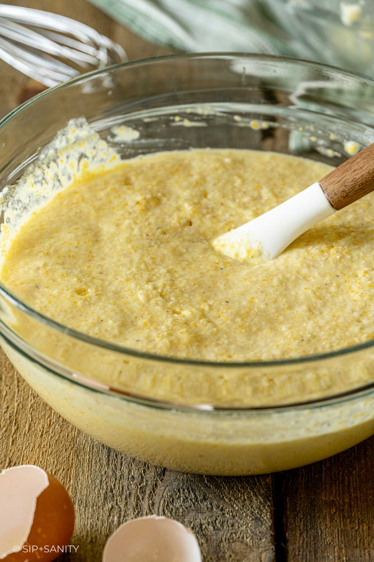 Glass bowl of yellow cornbread batter mixed with a white spatula beside eggshells and a metal whisk on wood.
