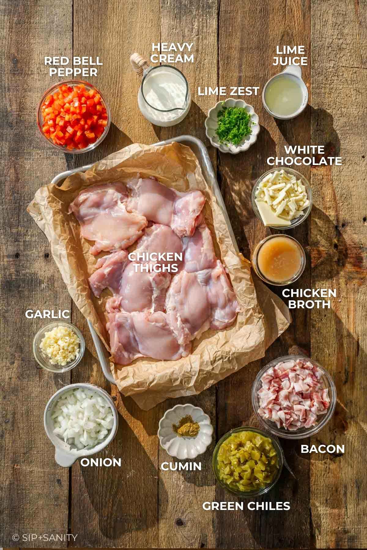 Raw chicken thighs on a tray, surrounded by bowls of peppers, cream, lime zest, broth, bacon, chiles, spices, onion.