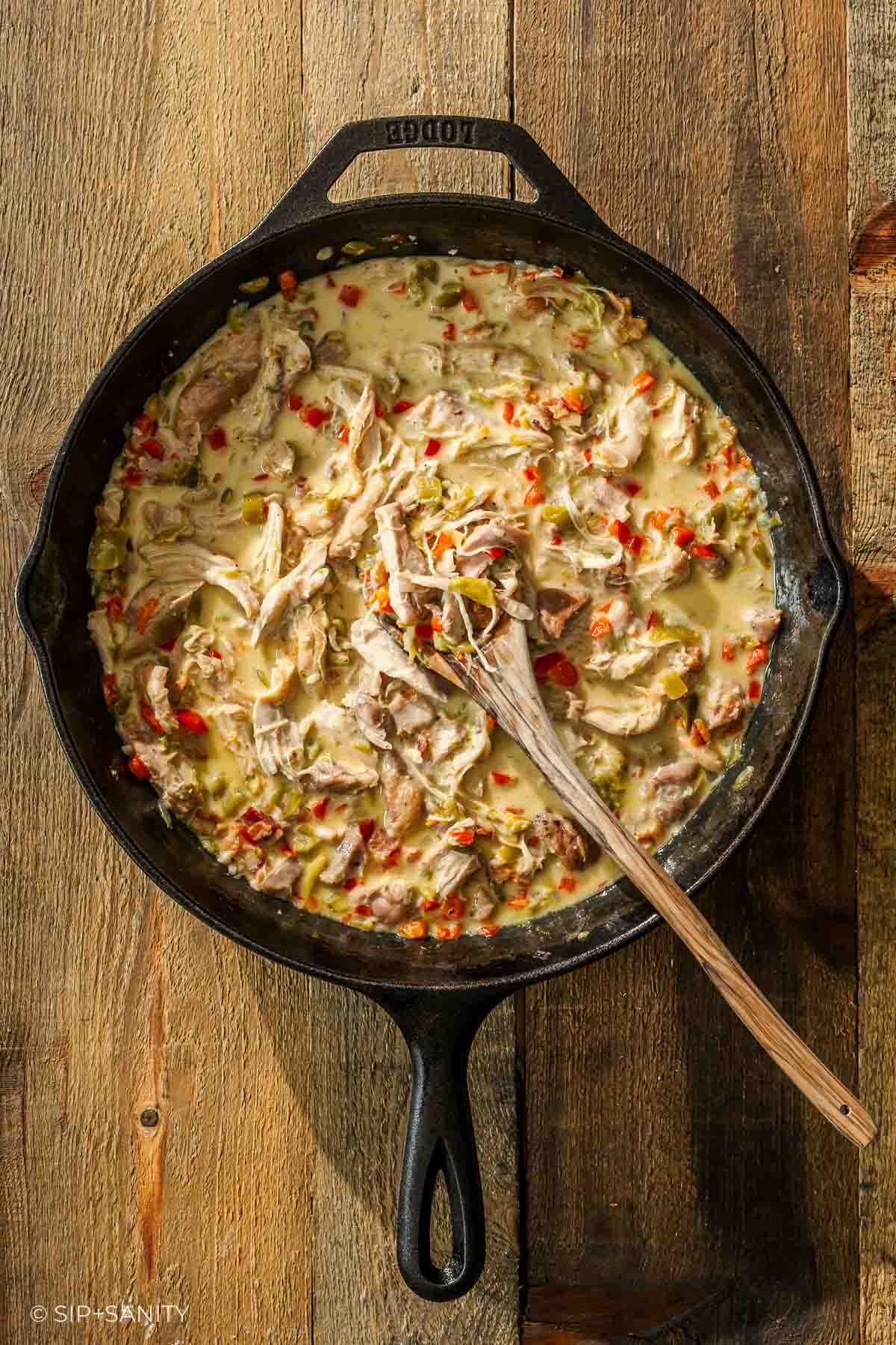 A black cast iron skillet of creamy chicken and red pepper vegetables sits on a wooden table with a spoon inside.