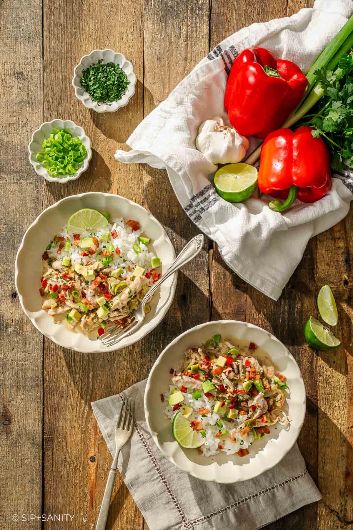Two bowls of rice with chicken, peppers, green onions, and lime on a rustic table with fresh veggies and utensils nearby.