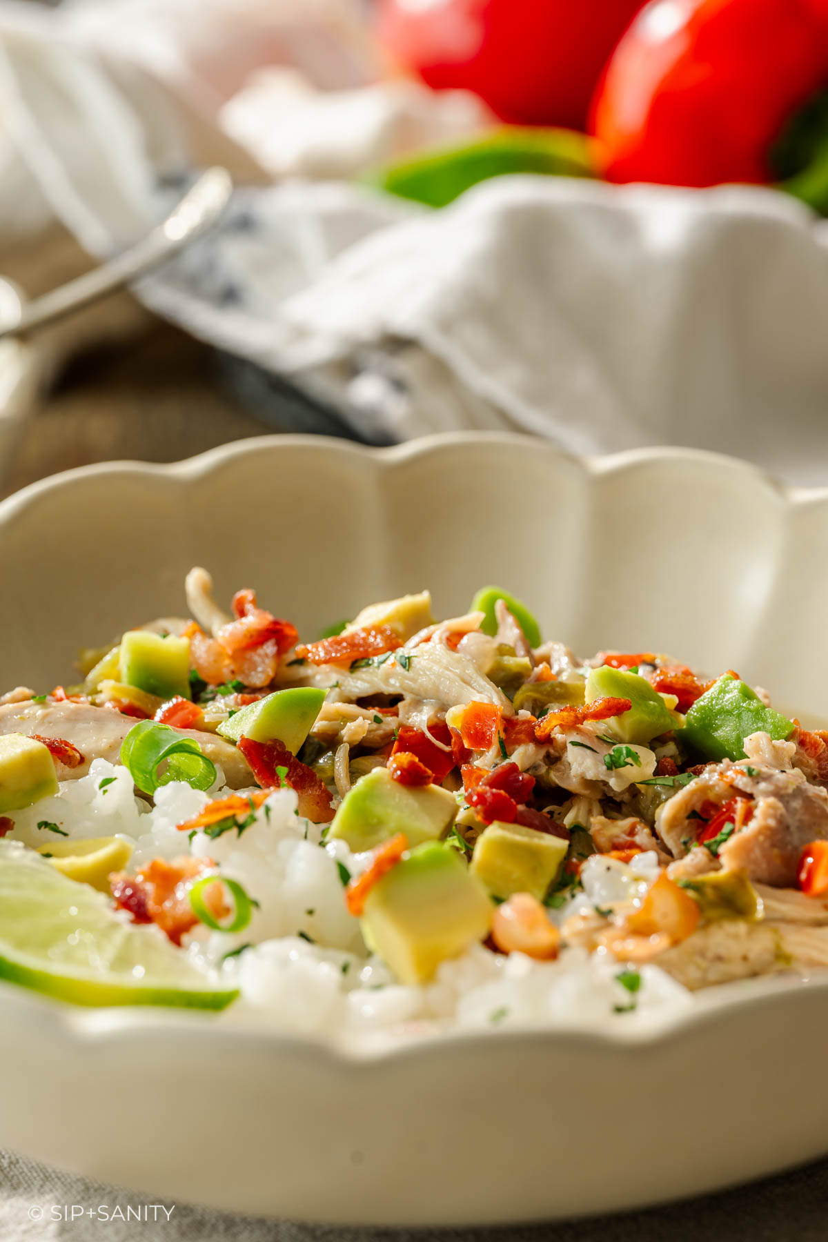 White rice in a bowl with avocado, green onions, chicken, bacon bits, and lime; tomatoes blurred in background.