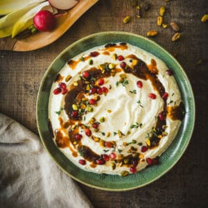Green bowl of creamy dip topped with pomegranate, pistachios, herbs, and sauce. Radishes and endive nearby.