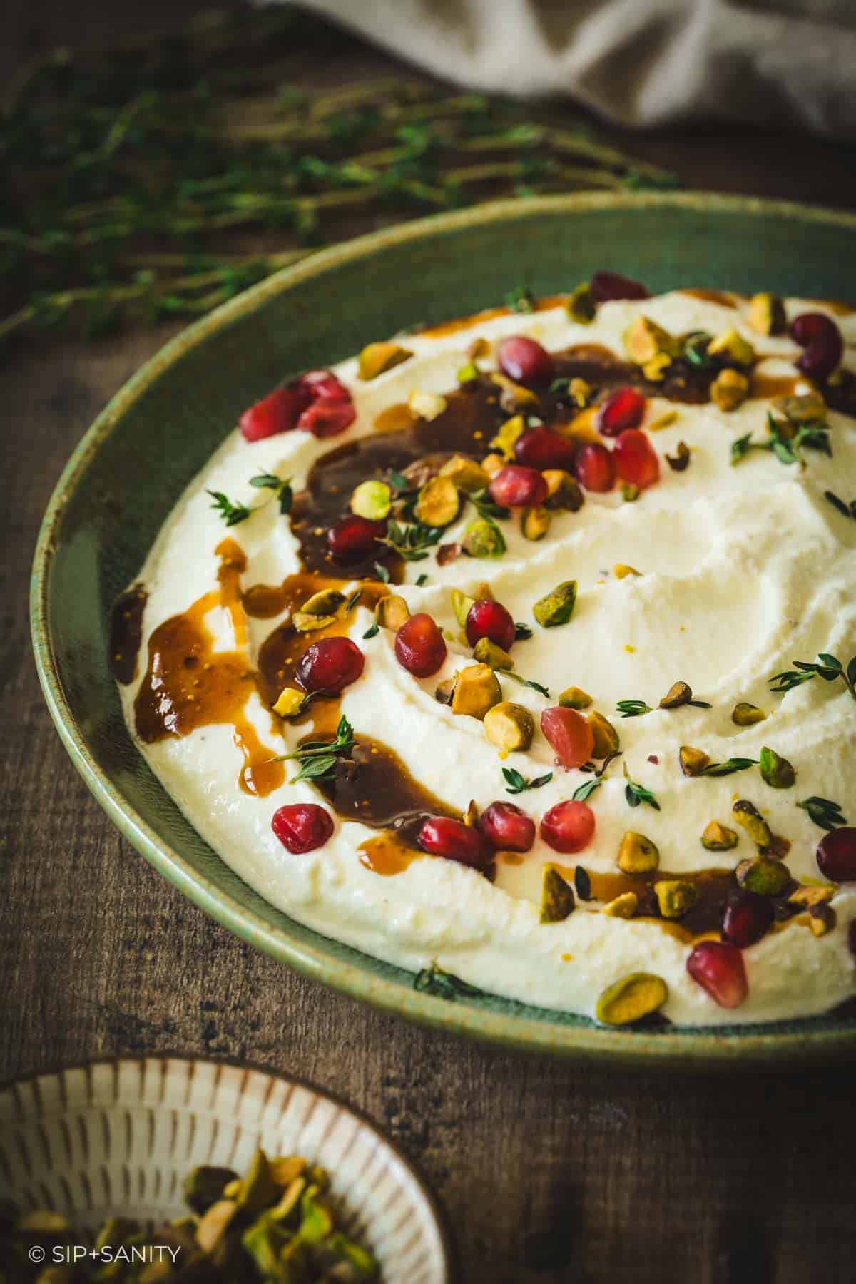 A green bowl of whipped ricotta-feta dip topped with pomegranate, pistachios, thyme, and dark sauce on a wood table.