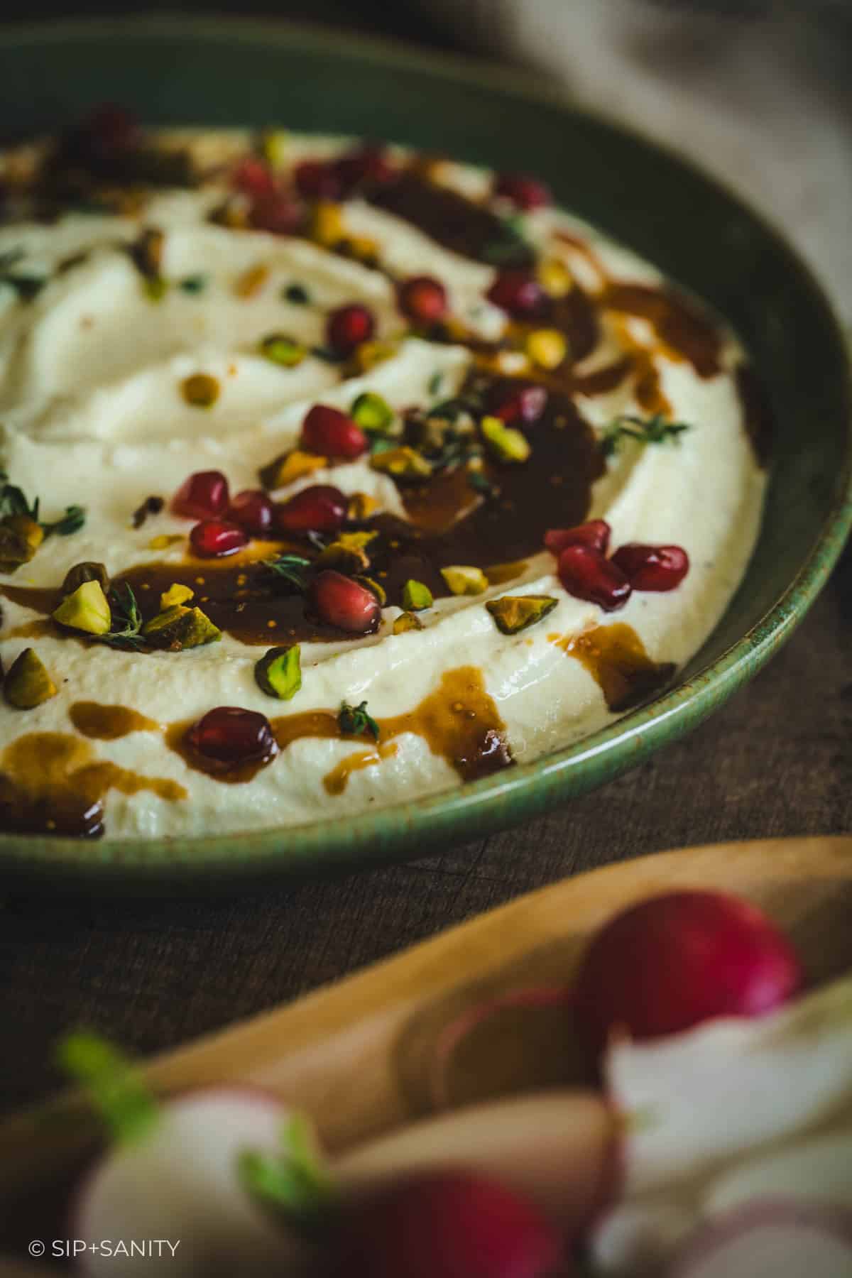 Whipped feta dip in a green bowl with pomegranate, pistachios, herbs, and sauce; radishes on a wooden plate nearby.