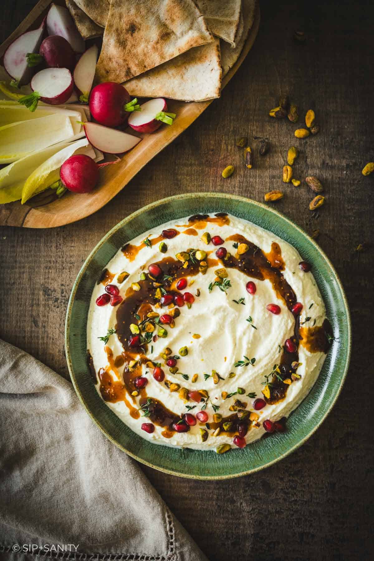 Whipped feta with herbs, pomegranate seeds, sauce in a bowl next to pita, endive, and radishes on a wooden table.