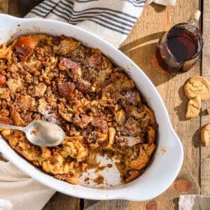 Gingerbread eggnog french toast in a white baking dish with a spoon, syrup, and cookies on a wooden table.