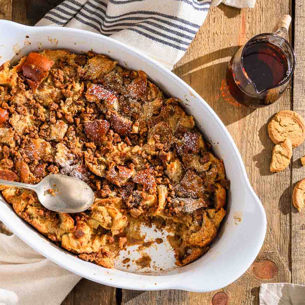 Gingerbread eggnog french toast in a white baking dish with a spoon, syrup, and cookies on a wooden table.