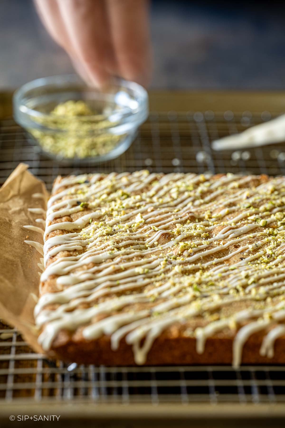 A square of eggnog blondie with white icing drizzle and chopped pistachios on a wire cooling rack.