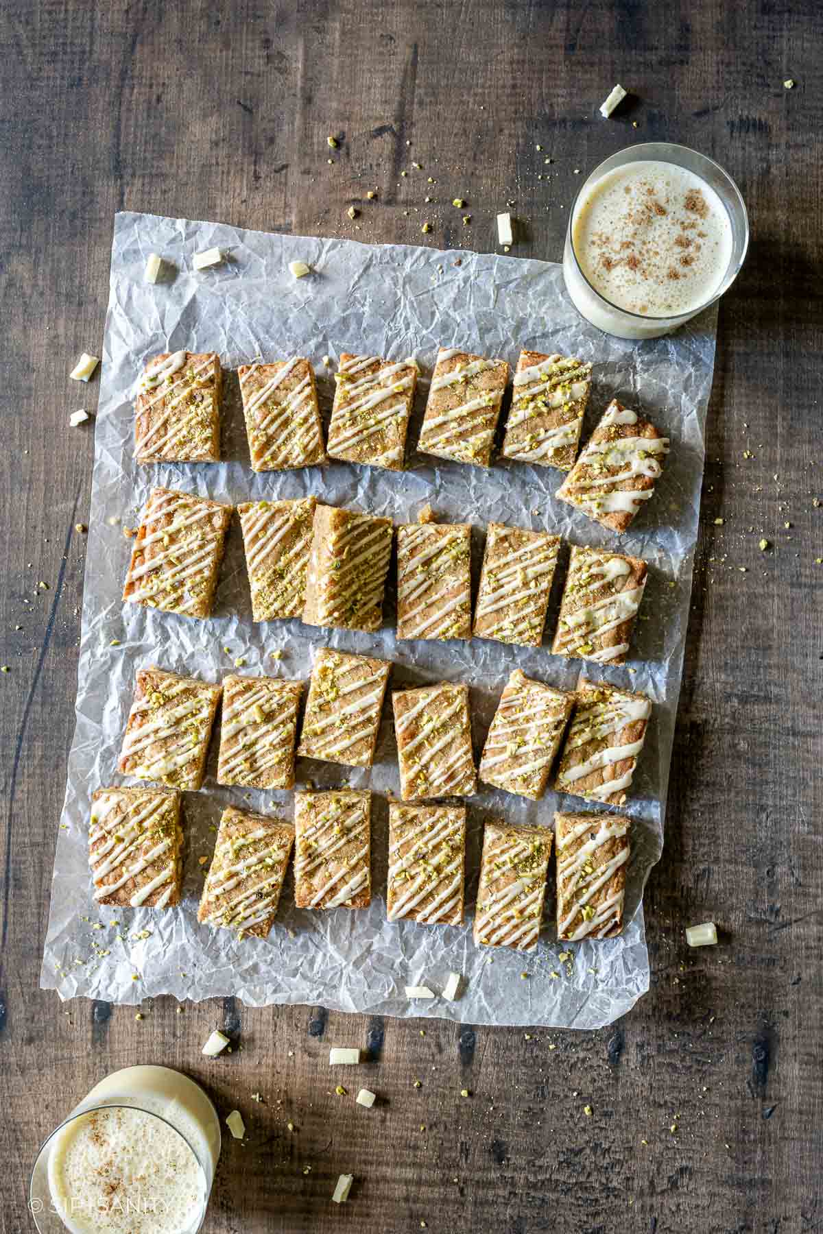 Rectangular eggnog blondie bars with white icing and nuts on parchment, paired with two glasses of eggnog.