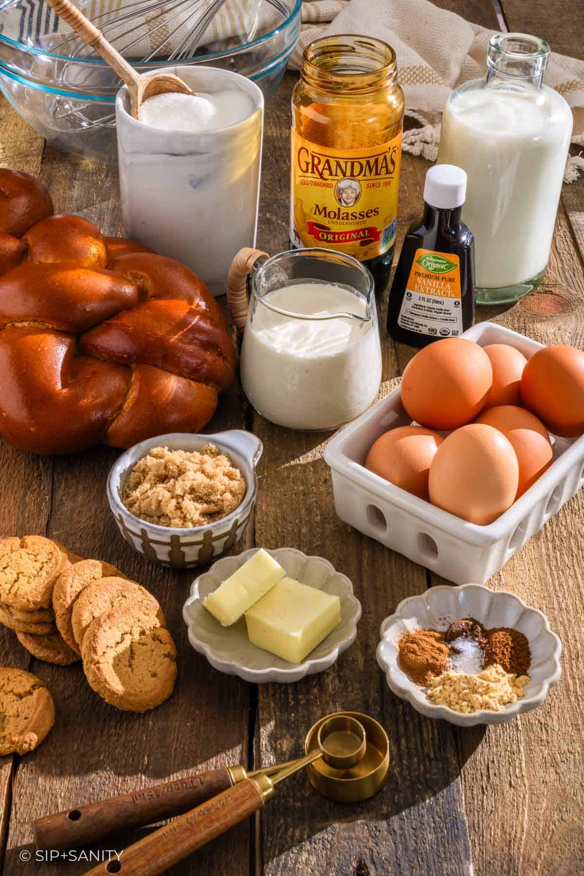 Baking ingredients on a wooden table: a braided loaf, eggs, molasses, milk, spices, and cookies.