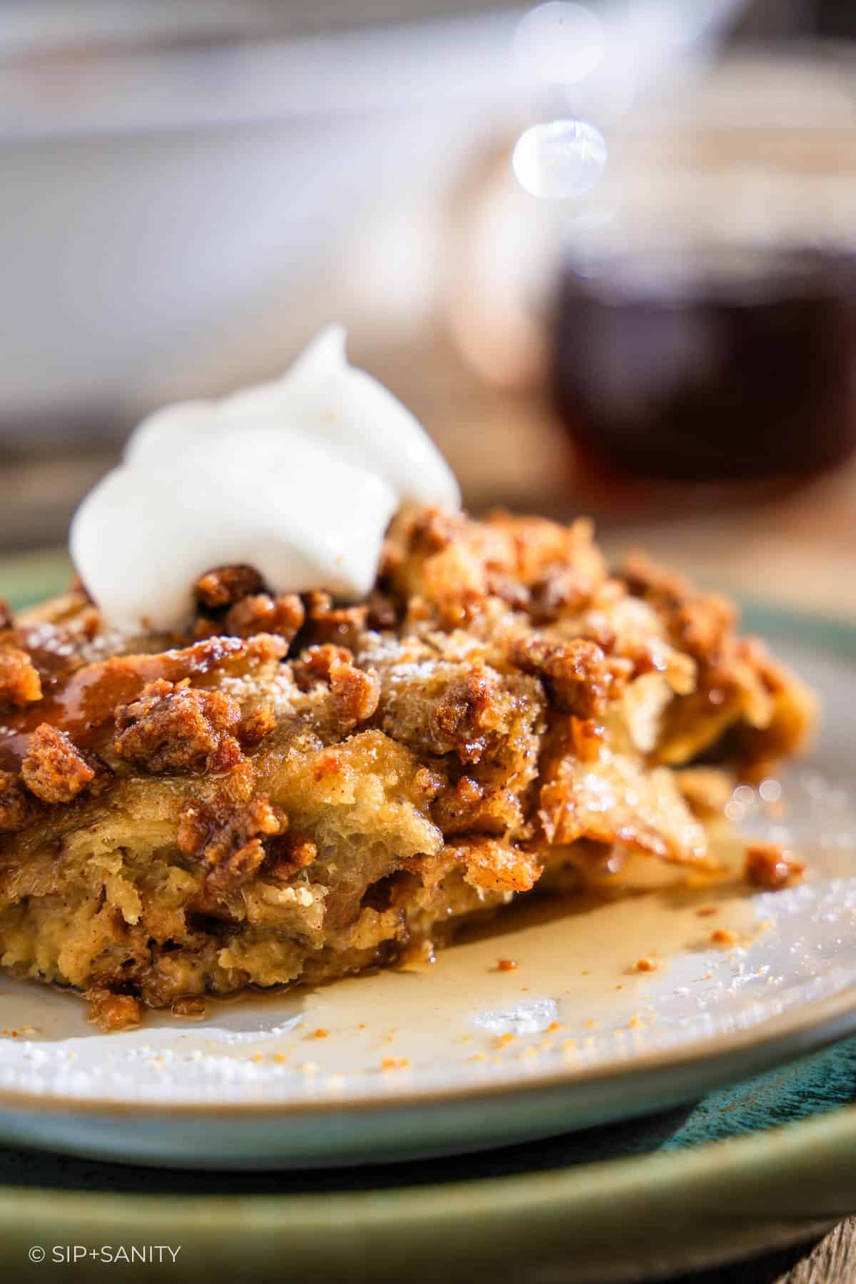 A square of gingerbread eggnog french toast casserole on a plate, topped with pecans, syrup, and whipped cream.