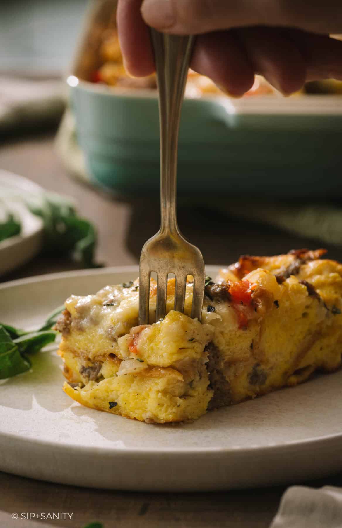 A fork lifts a bite of square egg and sausage breakfast casserole from a plate.